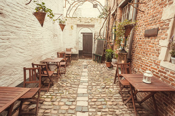 Empty space with wooden tables for hungry visitors of cozy outdoor restaurant in old style narrow street, with brick walls and cobbled stones