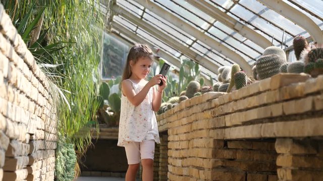 Child little girl takes mobile phone pictures of variety of cactus plants in a greenhouse of the botanical garden