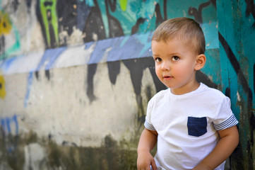 Fototapeta premium A two-year-old baby boy in hip hop style clothes sits under a wall with green graffiti and looks inquiringly upwards