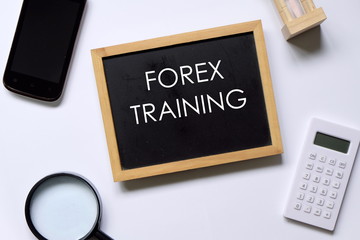 Top view of mobile phone,hourglass,calculator,magnifying glasses,and blackboard written with FOREX TRAINING on white background.