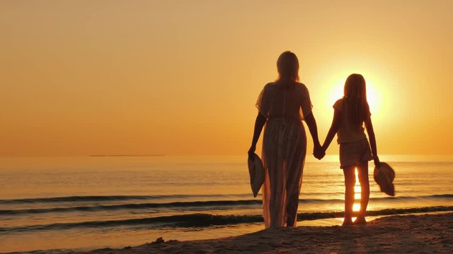 A woman holds a child's hand, stand together on the seashore, look at a beautiful sunset. Mother with daughter