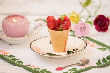 Dessert - red color sherbet ice cream in a waffle cone on the rose flowers saucer, cloth napkin next to a burning candle and three roses on the white table