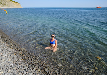 A beautiful girl in a blue bikini sits in sea water near a rocky beach. Transparent sea water.