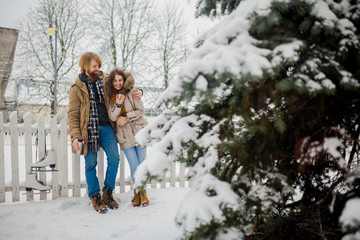 Fototapeta na wymiar Young loving couple caucasian man with blond long hair and beard, beautiful woman have fun drinking a hot drink from thermos, eating green apple in snow park near white fence and coniferous tree.