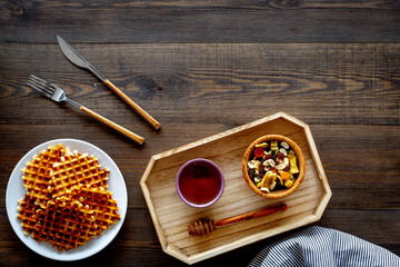 Round belgian waffles for breakfast. Breakfast in bed. Waffles on plate. Honey and dried fruits in tray, knife and fork, tablecloth on dark wooden background top view copy space
