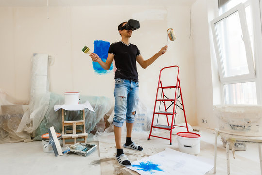 Young man with dark hair is painting the wall in white in his apartment. In his hands he holds a paint roller and paint brush. Can not decide what to choose.