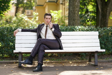 Obraz premium a young man in a business suit left the office and came to the Park. he sits on a white bench alone and talks on the phone
