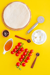 Ingredients for cooking pizza. Rolled out pizza dough, cherry tomatoes, olive oil, cheese mozzarella, spices on yellow background top view mockup