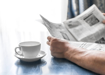 A man reading newspaper and drinking coffee sitting in cafe or at home next to kitchen window at sunny morning time. A coffee cup on dark blue table. A person hands holding newspaper