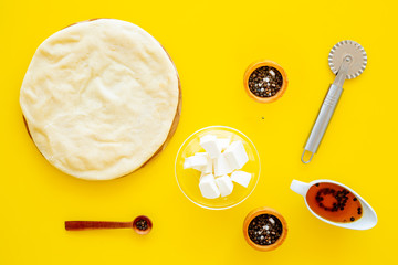 Raw rolled out pizza dough near cheese mozzarella, olive oil, spices, knife for pizza on yellow background top view mockup