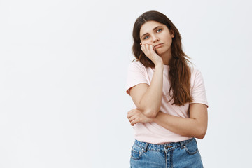 Studio shot of bored indifferent brunette woman with long hair in trendy outfit leaning head on hand and gazing with boredom and careless look at camera dreaming about summer vacation over gray wall