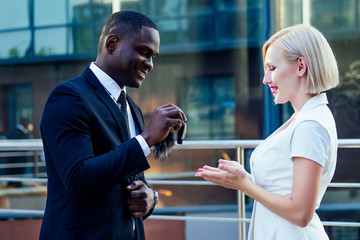 handsome Afro businessman in a stylish black suit gives car keys to attractive blonde business lady on street background office skyscraper. rent and purchase automobile
