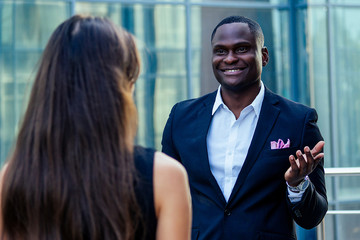 handsome Afro businessman in a stylish black suit looking talking with a woman on street background office skyscraper. luck and successful deal concept