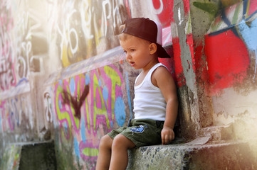 A two-year-old baby boy in a hip hop style clothes is sitting alone under the wall with graffiti and very upset