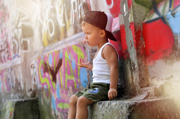 A two-year-old baby boy in a hip hop style clothes is sitting alone under the wall with graffiti and very upset