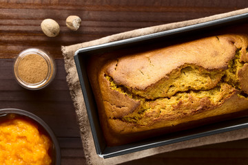 Fresh homemade pumpkin bread in pan, ingredients (pumpkin puree, cinnamon, nutmeg) on the side, photographed overhead (Selective Focus, Focus on the top of the bread)