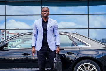 successful businessman handsome African American man in a stylish suit in a blue jacket standing in front of a cool new black car on the street