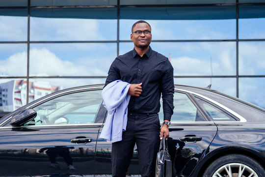 successful businessman handsome African American man in a stylish suit in a blue jacket black shirt and glasses standing in front of a cool new black car on the street