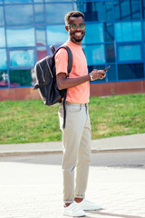 handsome trendy African Americans nerd male with glasses in stylish clothes colorful T-shirt with a backpack on the shoulders stand against the background of the blue windows of the university