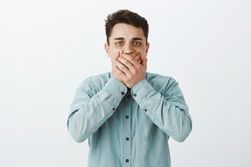 Studio shot of uncomfortable worried caucasian man in shirt, covering mouth with palms and staring at camera, wanting vomit or having stomach distorder, standing over gray background