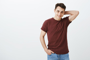 Studio shot of good-looking confident young man in red t-shirt, touching back of head and smiling playfully while flirting with woman in bar, asking number over gray background