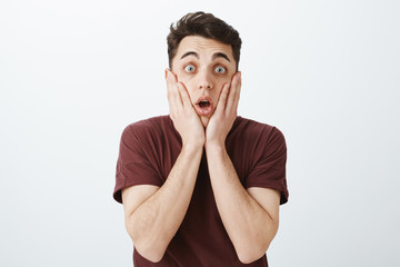 Indoor shot of shocked and stunned handsome man in red t-shirt, dropping jaw, gasping and holding...