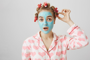 Funny white female in hair-curlers and pyjamas with heart print, standing in face mask and looking at camera, checking waverer on head and standing over gray background. Girl focused on taking it off