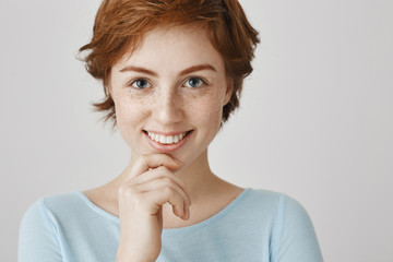 Fototapeta na wymiar Eyes are mirror of soul. Close-up portrait of attractive female with ginger hair and freckles holding hand on chin and smiling broadly, gazing at camera with interested expression over gray wall