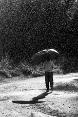 A small unidentified boy walks in the rain in Finland. He has a black umbrella. Image has a black and white effect.