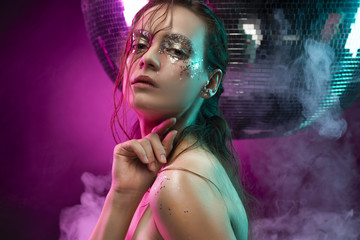 Beautiful girl with creative makeup made of glitter with tears on her face illuminated with pink and blue light in a scenic smoke near the big mirror ball on pink