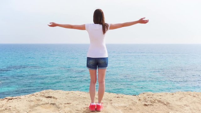 Smiling young woman in white t-shirt raising arms up at cliff against breathtaking view of blue sea. Carefree female outstretching hands in slow motion. Concept of freedom and inspiration
