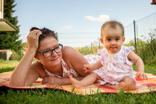 Adorable infant baby girl with mother on colorful blanket in green grass. Ninth months old child having fun with mum in summer garden.