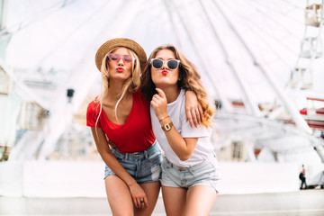 Best friend having fun near Ferris wheel, going crazy together, wearing hats and mirrored sunglasses, amazing view on the city, bright colors evening sunlight