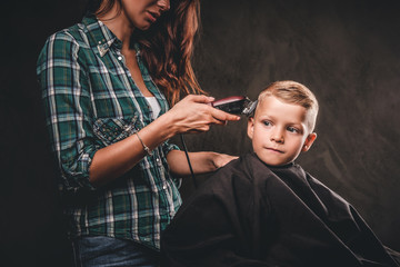 Obraz premium Children hairdresser with the trimmer is cutting little boy against the dark background. Cute preschooler boy getting haircut. 