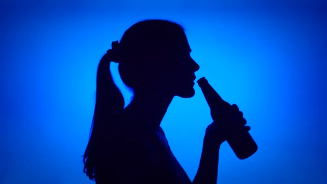 Silhouette of young woman opening can beer bottle on blue background. Female's face in profile drinking beer from glass bottle. Black contur shadow of teenager's half-face