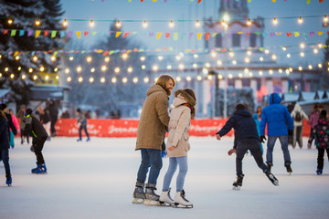 Young couple in love Caucasian man with blond hair with long hair and beard and beautiful woman have fun, active date skating on ice scene in town square in winter on Christmas Eve