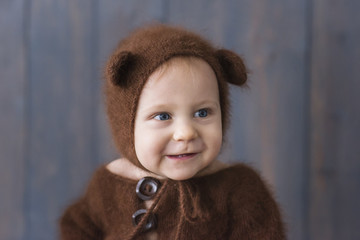 Close-up portrait of a little boy in a brown, fluffy, knitted bear costume, sitting on a wood floor, playing with a bright Christmas garland of lights. The kid laughs, sticks out his tongue. New Year