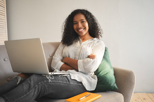 Candid shot of happy successful young dark skinned female blogger sitting on sofa with modern electronic device on her lap, keeping arms crossed and smiling confidently at camera, browsing internet