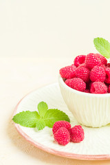 Ripe raspberries with green mint leaves in cup and saucer on pastel yellow background.