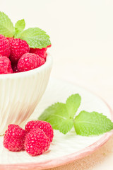 Ripe raspberries with green mint leaves in cup and saucer on pastel yellow background.