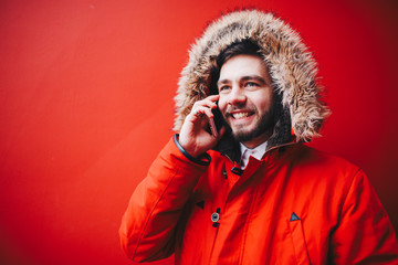 handsome young male student with a toothy smile and a beard stands on a red wall background in a bright red winter jacket with a hood with fur in winter. Uses a mobile phone, talk and call