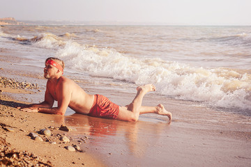 guy in red swimming trunks and a bandana on his head, lies on a sandy beach and is covered by a...