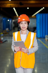 Portrait of female factory worker wearing hardhat and reflective vest looking at camera and smiling cheerfully while posing in workshop of production plant, copy space