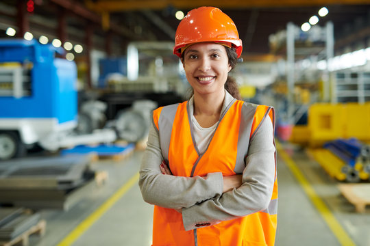Waist up portrait of cheerful young woman wearing hardhat smiling happily looking at camera while posing confidently in production workshop, copy space