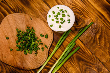 Glass bowl with sour cream and cutting board with chopped green onion on wooden table. Top view