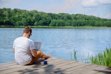Fototapeta na wymiar man working on laptop while sitting on wooden dock. legs in river water