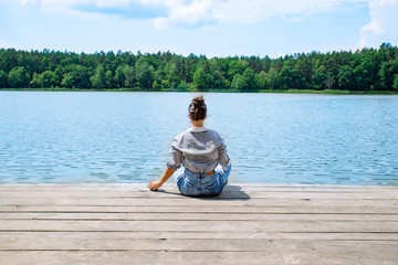 woman sitting on wooden dock looking at lake in sunny day - obrazy, fototapety, plakaty