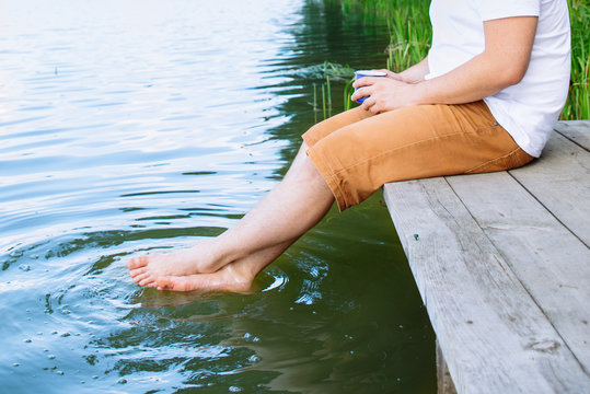 Man Close Up Sitting At Wooden Pier Legs In Water. Drinking Coffee. Crop