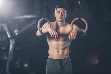 Brooding man is standing and leaning on sport rings. He is resting after acrobatic training with...