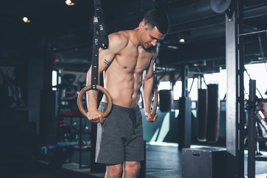 Shirtless guy is doing hard support exercise with equipment. He is pulling up while holding rings and balancing in this position. Male is having workout in gym